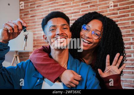 Das junge afroamerikanische Paar zeigt die Schlüssel ihres ersten Hauses nach dem Kauf und zieht zusammen in ein neues Zuhause. Glückliche Ehemann und Frau, die sich in ihrer Wohnung umarmen und sich freuen, Besitzer einer Wohnung zu sein. Hochwertige Fotos Stockfoto