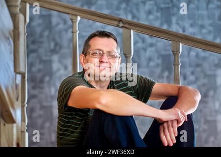 Ptosis des linken Lids bei reifem Europäermann, der zu Hause auf einer Holzzwischenflurtreppe sitzt. Stockfoto