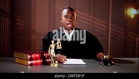 Selbstbewusster Reifer Männlicher Richter Schlägt Den Gavel Am Tisch Gegen Bücherregal Stockfoto
