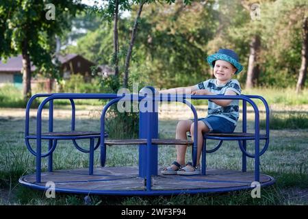 Ein vierjähriger Junge in blauer Kleidung dreht sich auf dem Spielplatzkarussell. Stockfoto