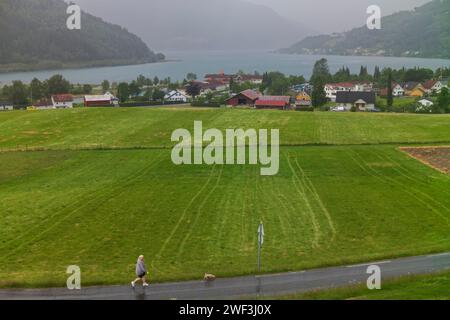 Loen, Norwegen, 26. Juni 2023: Eine Frau geht mit ihrem Hund während eines regnerischen Tages im Dorf Loen, Norwegen, fotografiert von einem Felsvorsprung, der den Schlepper überblickt Stockfoto