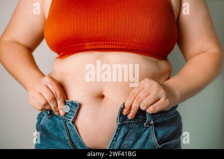 Frauen Körperfettbauch. Übergewichtige Frauenhand mit übermäßigem Bauchfett. Eine fette Frau, die versucht, ihre Jeanshose zu ziehen. Frauengesundheit. Stockfoto