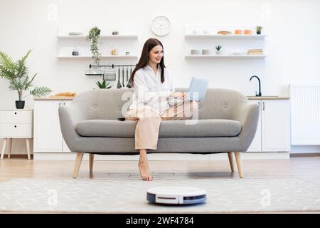 Junge kaukasische Frau überwacht Roboter-Staubsauger auf einem persönlichen Laptop, während sie mit überkreuzten Beinen auf dem Sofa in einer hellen Küche sitzt. Einfache Reinigung mit Stockfoto