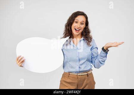 Eine fröhliche Frau mit lockigem Haar und einem blauen Hemd hält ein weißes Sprechblase-Zeichen Stockfoto