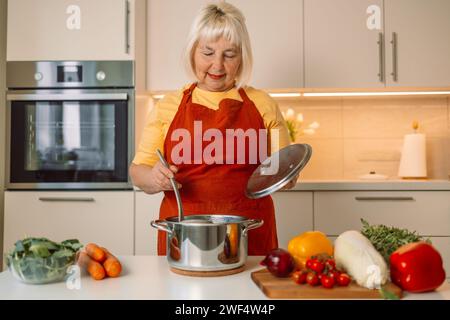 Ältere Frau aus den 60er Jahren, die in weißer Küche steht und riecht, frisch köstlich von Suppe in einem Topf mit Dampf schmeckt Stockfoto