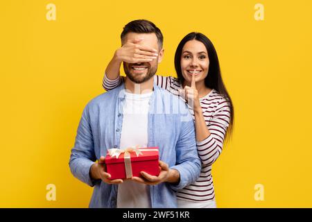 Eine liebende Frau, die die Augen des Mannes bedeckt und ihn mit einem Geschenk überrascht Stockfoto