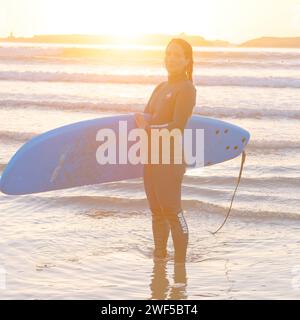 Eine Surferin lächelt im Neoprenanzug und hält ihr Surfbrett bei Sonnenuntergang in Essaouira, Marokko, 28. Januar 2024 Stockfoto