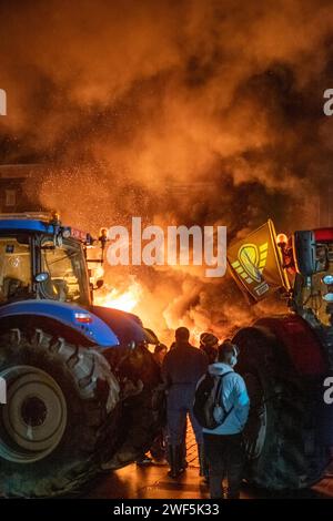 Turnhout, Antwerpen, Belgien, 28. Januar 2024, das Bild zeigt eine eindrucksvolle Szene aus dem Protest der Bauern gegen das Stickstoffabkommen in Turnhout, Belgien. Mitten in der Nacht bildet das glühende Licht eines Lagerfeuers die Bühne, mit dickem Rauch, der in den dunklen Himmel strömt. Der Traktor, ein Symbol des landwirtschaftlichen Berufs, ist an prominenter Stelle positioniert, während sich eine Gruppe von Demonstranten sammelt, die einige Flaggen halten, die für ihre Sache repräsentativ sind. Die intensive und temperamentvolle Atmosphäre ist spürbar, wobei die dramatische Beleuchtung die kritische Natur des Protestes widerspiegelt. Nachtwache von Bauernprotest i Stockfoto