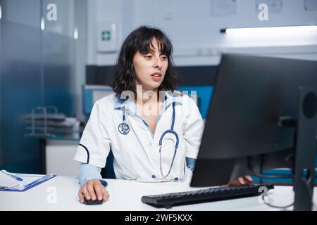 Fokussierte Ärztin mit einem Desktop-pc in einem Krankenhausbüro. In einem Klinikzimmer, ein kaukasischer Arzt, der einen Laborkittel trägt und einen Computer benutzt, um medizinische Akten zu überprüfen. Stockfoto