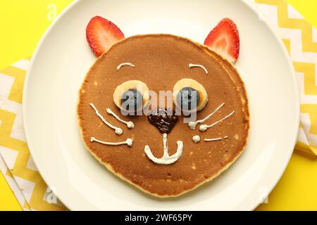 Kreatives Servieren für Kinder. Teller mit niedlicher Katze aus Pfannkuchen, Beeren, Creme, Banane und Schokoladenpaste auf gelbem Hintergrund, Draufsicht Stockfoto