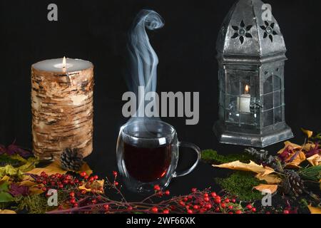 Dampfende Tasse Tee umgeben von Herbstblättern und Laub, brennender Kerze und Laterne vor schwarzem Hintergrund Stockfoto