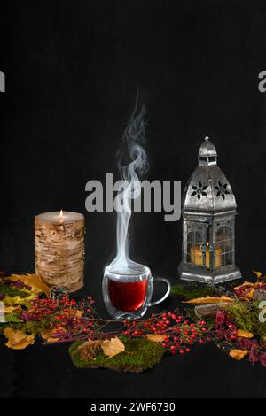 Dampfende Tasse Tee umgeben von Herbstblättern und Laub, brennender Kerze und Laterne vor schwarzem Hintergrund Stockfoto