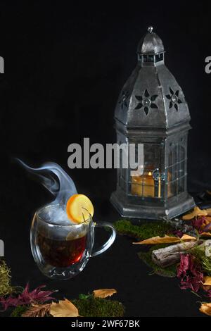Dampfende Tasse Tee umgeben von Herbstblättern und Laub, brennende Kerze in Laterne vor schwarzem Hintergrund Stockfoto
