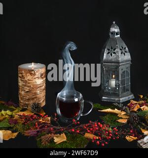 Dampfende Tasse Tee umgeben von Herbstblättern und Laub, brennender Kerze und Laterne vor schwarzem Hintergrund Stockfoto