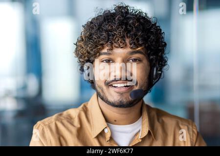 Porträt eines positiven indianers, der ein Headset mit Mikrofon trägt und die Kamera auf einem unscharfen Hintergrund betrachtet. Freundlicher Kundendienstmitarbeiter, der den Kunden berät und Probleme im Callcenter löst. Stockfoto