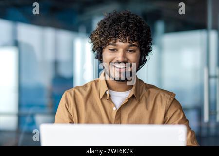 Lächelnder arabischer Mann mit lockigen Haaren im schnurlosen Headset, der im Büro auf den Laptop-Bildschirm schaut. Zufriedener Programmierer, der in der Ausbildung arbeitet, sieht gerne ein lehrreiches Webinar an, um die Produktivität zu steigern. Stockfoto