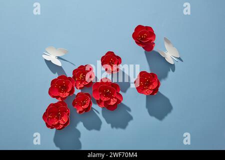 Kreative Layout-Papierkarte mit roten Papierblumen und weißem Schmetterling auf blauem Hintergrund. Frühlingsblumen-Konzeptrahmen für Werbung. Draufsicht, flache La Stockfoto