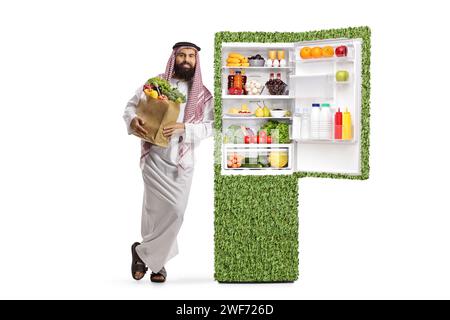 Saudischer arabischer Mann in ethnischen Kleidern, der neben einem umweltfreundlichen grünen Kühlschrank steht, der auf weißem Hintergrund isoliert ist Stockfoto