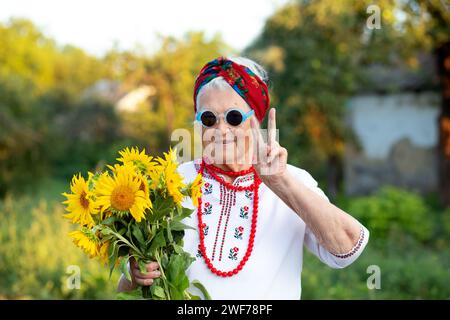 Gelb blaues Herz und ein Blumenstrauß in den Händen einer alten Großmutter in einem gestickten Hemd und roten Perlen. Zeigt die Geste von V Stockfoto