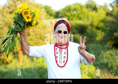 Sonnenblumenstrauß in den Händen einer alten Großmutter in einem gestickten Hemd und roten Perlen. Zeigt die Geste des Sieges Unabhängigkeitstag Stockfoto