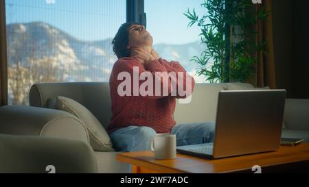 Müder junger Mann, der auf der Couch sitzt, hat starke Nackenschmerzen, reibt ihn, um Muskelverspannungen zu lösen, massiert seinen Hals und macht eine Pause von der Wor Stockfoto