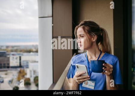 Weibliche Krankenschwester genießt nach der Arbeit eine Tasse Kaffee zu Hause und steht am Fenster. Vormittagstee vor der Arbeit. Work-Life-Balance für das Pflegepersonal. Stockfoto