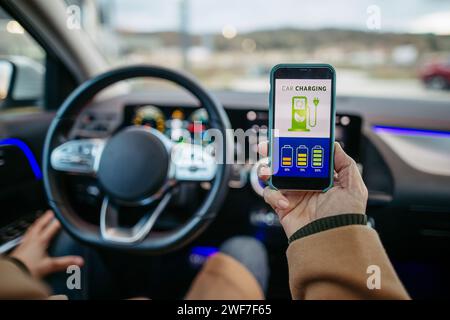Nahaufnahme eines Mannes, der die App zum Laden eines Elektrofahrzeugs verwendet und den Akkuladestand auf dem Smartphone überprüft. Laden von Apps zur Überwachung des Stromverbrauchs, Lokalisierung Stockfoto