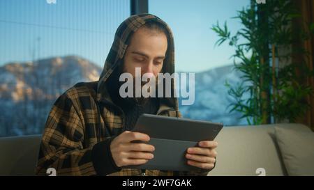 Bärtiger junger Mann mit Hoodie, der Videospiele auf einem Tablet-Computer spielt, während er zu Hause auf dem Sofa sitzt. Aufgeregt konzentrierter Mann-Engros Stockfoto