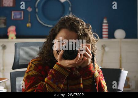 Junge weiße Frau mit lockigem Haar, die mit einer spiegellosen sony Alpha 7 II Kamera in einem Heimbüro mit blauer und weißer Wand ein Foto von sich selbst macht Stockfoto