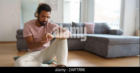 Junger Mann, der sich nach dem Heimtraining ausruht und seine Trainingsleistung auf Smartwatch überprüft. Morgens oder abends trainieren und Musik hören Stockfoto