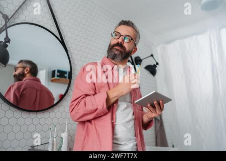 Ein Mann, der online einkaufen und sich im Badezimmer die Zähne putzt. Tablette halten, scrollen und kaufen, Zahnbürste im Mund. Stockfoto