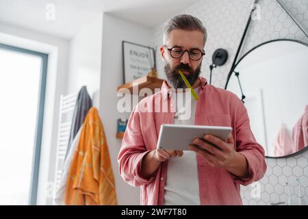 Ein Mann, der online einkaufen und sich im Badezimmer die Zähne putzt. Tablette halten, scrollen und kaufen, Zahnbürste im Mund. Stockfoto