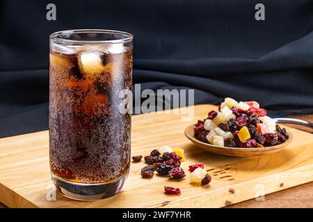 Nahaufnahme von kaltem Cola-Wasser mit Eiswürfeln in transparentem Glas und verschiedenen getrockneten tropischen Früchten in Holzschale auf Holzschneidebrett auf schwarzem Backgr Stockfoto