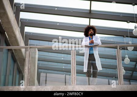 Porträt eines Arztes, der im Krankenhaus steht, sich auf Glasgeländer stützt und in die Kamera blickt. Stockfoto