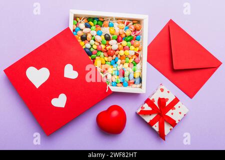 Roter Umschlag mit Süßigkeiten und Geschenkbox und Valentinsherzen auf farbigem Hintergrund. Flach liegend, Draufsicht. Romantischer Liebesbrief für das Urlaubskonzept. Stockfoto