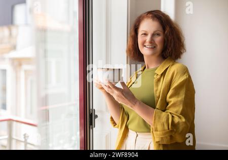 Schöne Seniorin genießt eine Tasse Kaffee, während sie am Fenster steht Stockfoto