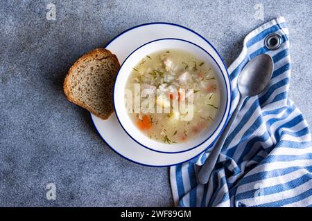 Blick von oben auf eine Schüssel mit Gemüse- und Hühnersuppe, begleitet von einem Stück Brot auf einer grauen Oberfläche mit einer blau-weiß gestreiften Serviette und einem spo Stockfoto