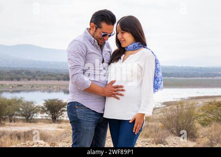 Ein Paar umarmt sich, während er in einer natürlichen Landschaft steht, während der Mann seine Hände auf den Bauch der schwangeren Frau legt und die Freude zeigt, die nat hervorhebt Stockfoto