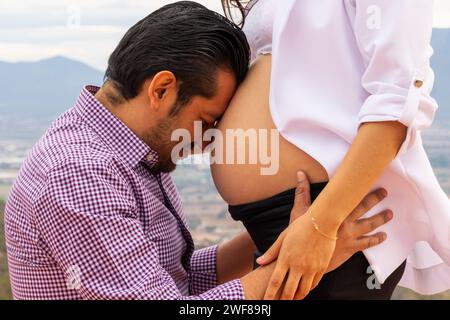 Eine intime Nahaufnahme eines Mannes, der den nackten Bauch seiner siebenmonatigen schwangeren Partnerin küsst, der seine Liebe mit einem malerischen Blick auf Tequisquiapan Mexiko ausdrückt Stockfoto