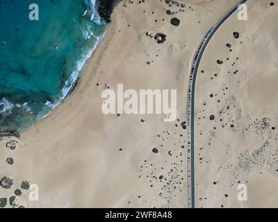 Aus der Vogelperspektive einer einzigen kurvenreichen Straße, die durch die weitläufigen Dünen von Corralejo führt, wobei der azurblaue Ozean sanft über die Küste von Fuerteventura ragt Stockfoto