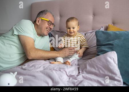 Fröhlicher älterer Mann lächelt, während er mit seinem kleinen Enkel auf einem Bett spielt, umgeben von weichen Kissen Stockfoto