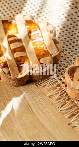 Ein sonnendurchflutetes italienisches Pastiera Napoletana wirft verspielte Schatten auf eine Holzfläche, umgeben von Osterelementen. Stockfoto