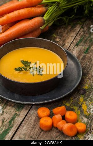 Rustikale und hausgemachte Karottensuppe, serviert in einer dunklen Schüssel, garniert mit Petersilie, zusammen mit frischen Karotten mit Grün auf verwittertem Holzhintergrund. Stockfoto