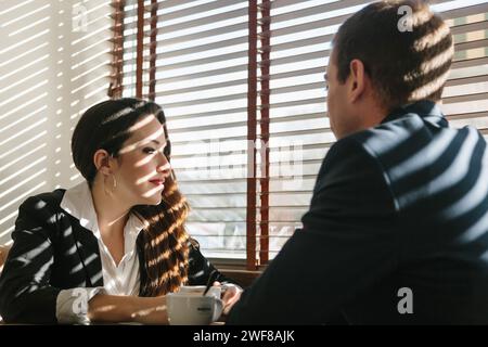 Zwei Fachleute führen in einem sonnendurchfluteten Café in Madrid eine konzentrierte Diskussion, während Schatten von den Jalousien über sie werfen. Stockfoto