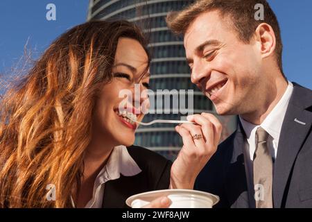 Ein Mann und eine Frau in Geschäftskleidung teilen einen unbeschwerten Moment mit einem einzigen Spaghetti-Strang vor dem Hintergrund der modernen Architektur Madrids. Stockfoto