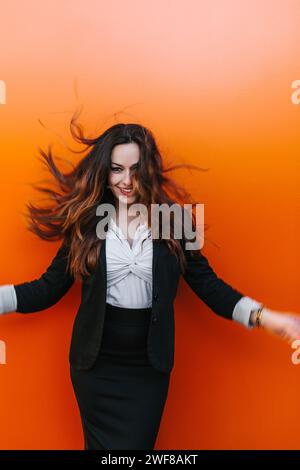 Eine lächelnde Frau, die in professioneller Kleidung in die Kamera schaut, schwingt ihr Haar gegen eine leuchtend orange Wand und hält einen Moment unbeschwerten Glücks in Ma fest Stockfoto