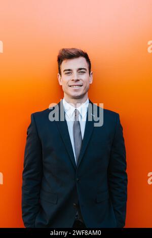 Ein junger, männlicher Profi, der die Kamera ansieht, steht in einem klassischen schwarzen Anzug mit Krawatte vor einer leuchtend orangefarbenen Wand in Madrid, Spanien. Stockfoto