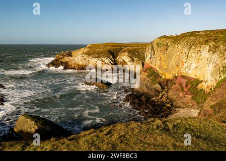 Blick hinunter auf die roten Felsen rund um Porth Saint Rhoscolyn vom Isle of Anglesey Coastal Path mit Wellen, die an einem windigen Tag an der Küste stürzen Stockfoto