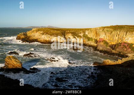 Blick hinunter auf die roten Felsen rund um Porth Saint Rhoscolyn vom Isle of Anglesey Coastal Path mit Wellen, die an einem windigen Tag an der Küste stürzen Stockfoto