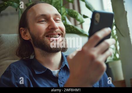 Entzückender bärtiger junger Mann mit Zahnspange, der mit Telefon in einem bequemen Sessel sitzt und in den sozialen Medien scrollen und lächelt Stockfoto